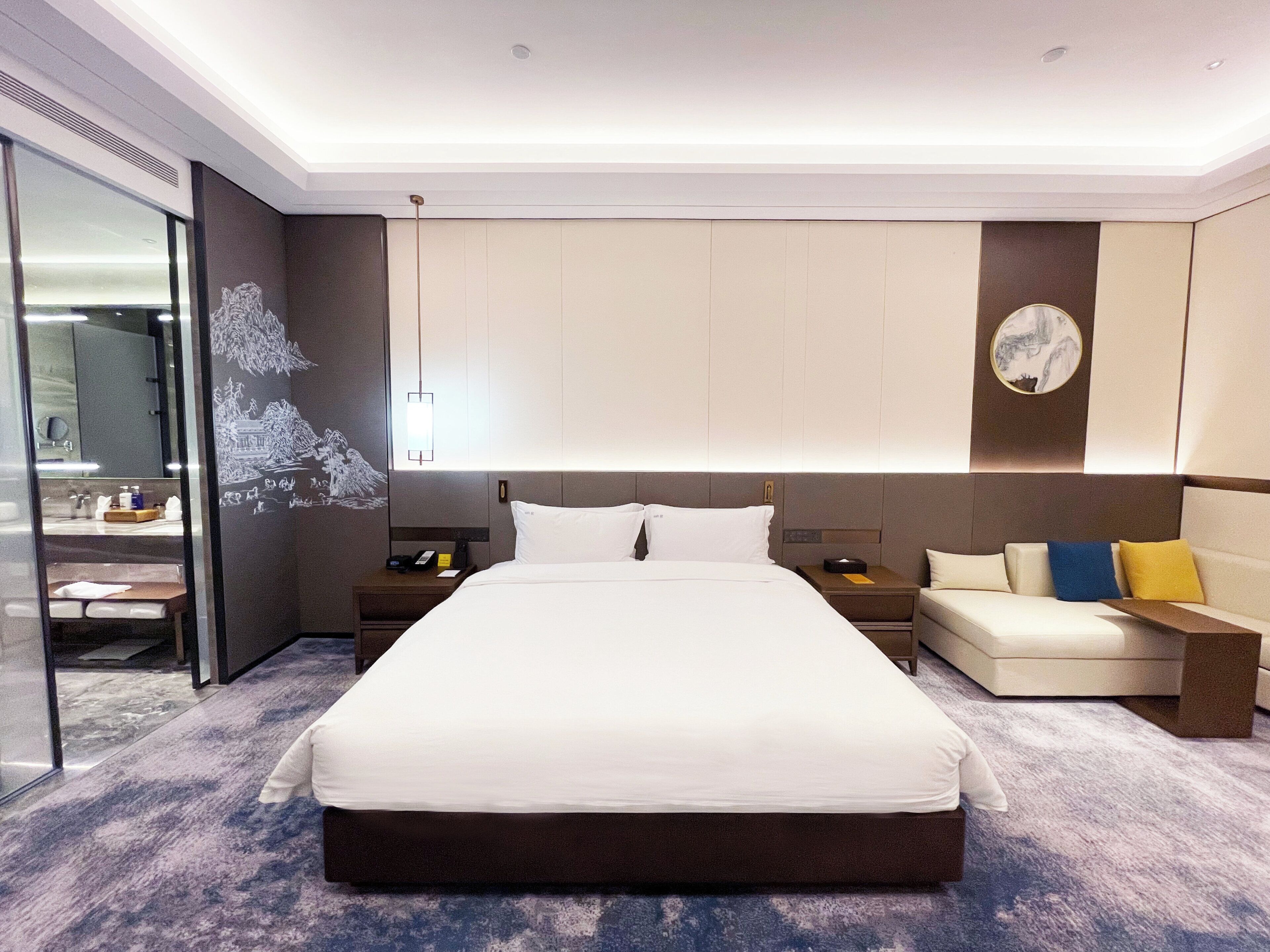 A modern hotel room with a large bed, a bedside table with a lamp, a sofa with cushions, and a large mirror.