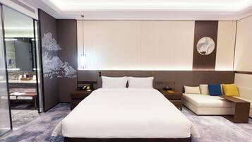 A modern hotel room with a large bed, a bedside table with a lamp, a sofa with cushions, and a large mirror.