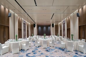 Uma sala de banquetes com mesas redondas montadas para refeições, cadeiras brancas e um tapete azul estampado.