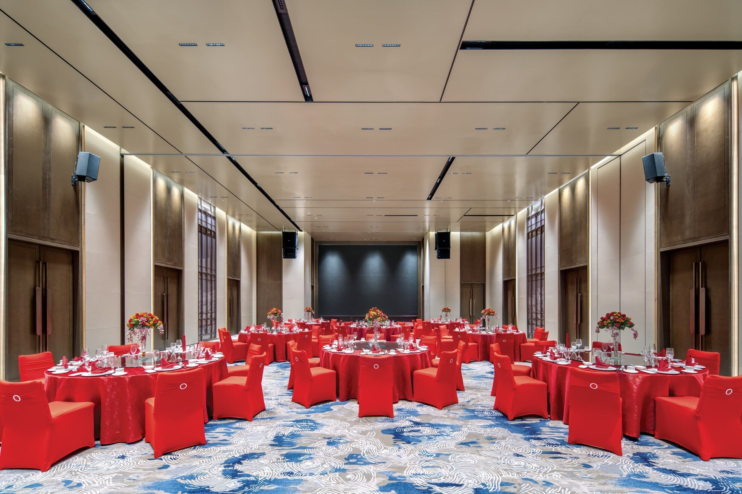 A large conference room with rows of round tables, red chairs, and a blue patterned carpet.