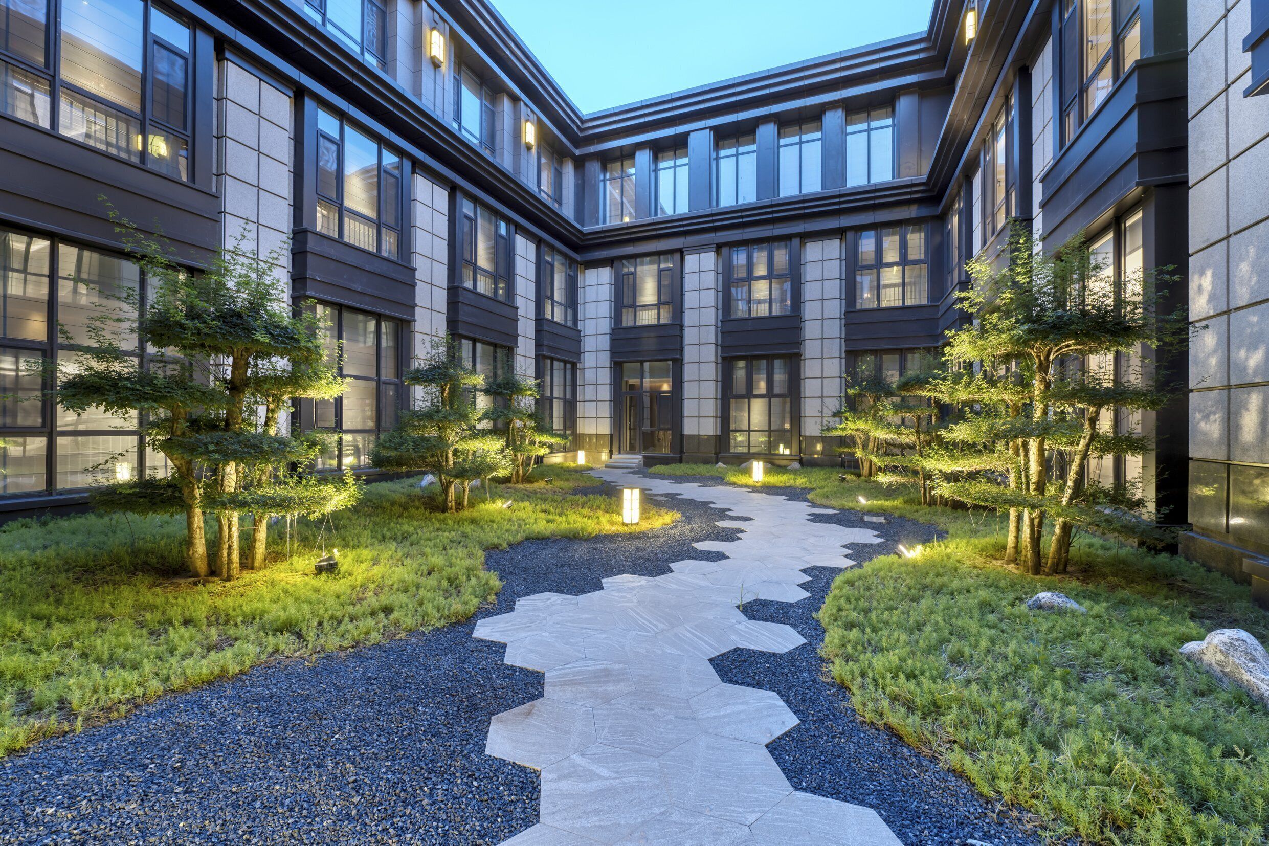 Un edificio moderno con un cortile centrale caratterizzato da un viale in pietra, circondato da alberi e illuminato da luci da terra.