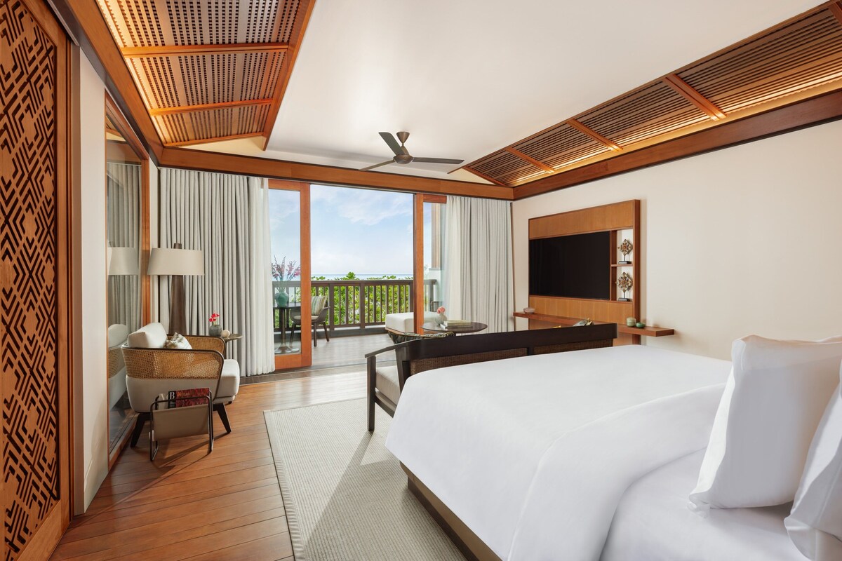 A hotel room with a large bed, a desk, a chair, a TV, and a balcony with a view.