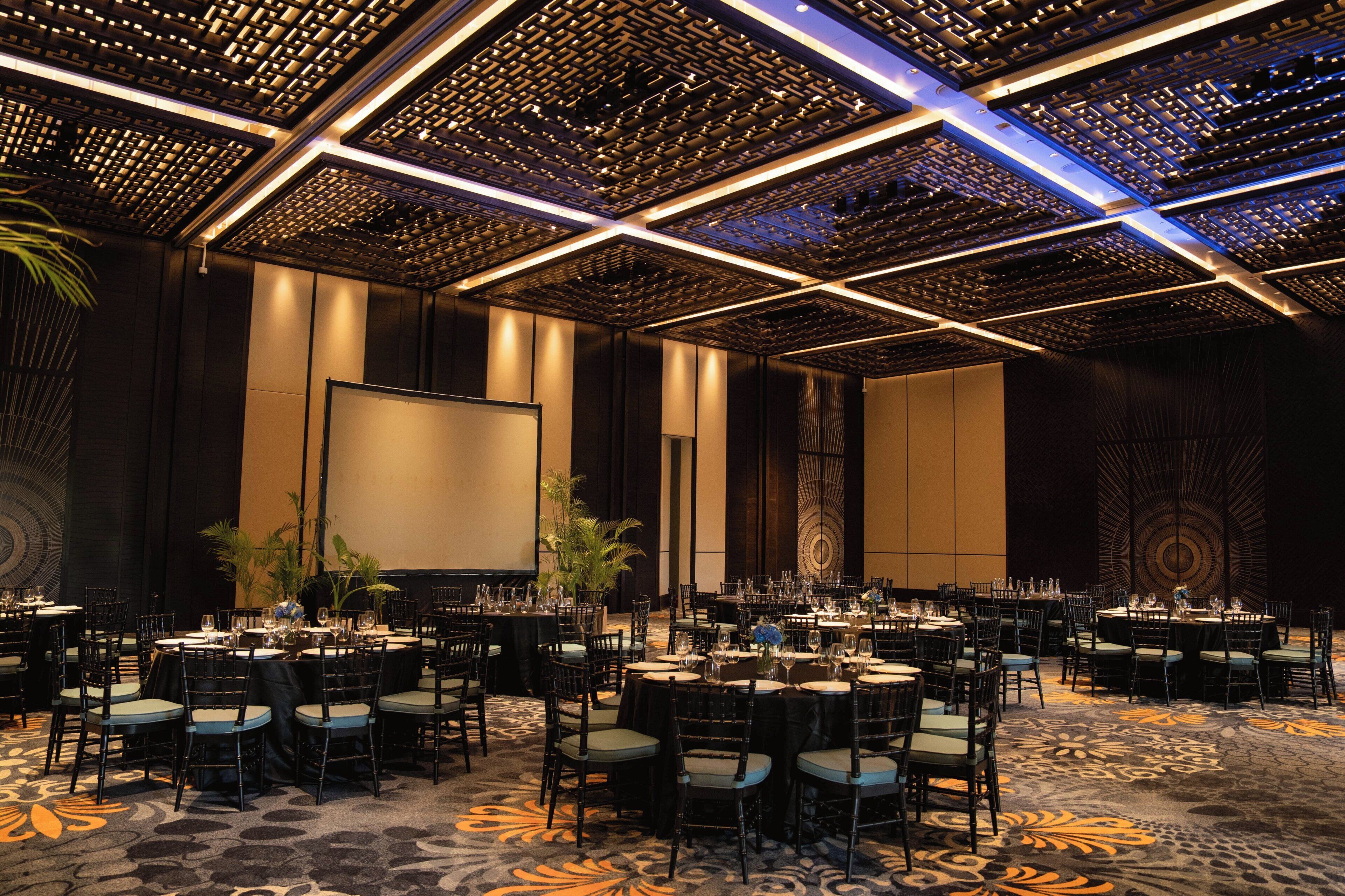 A banquet hall with round tables, chairs, and a large screen.