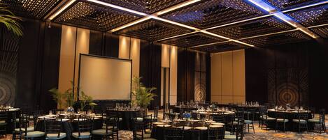 A banquet hall with round tables, chairs, and a large screen.