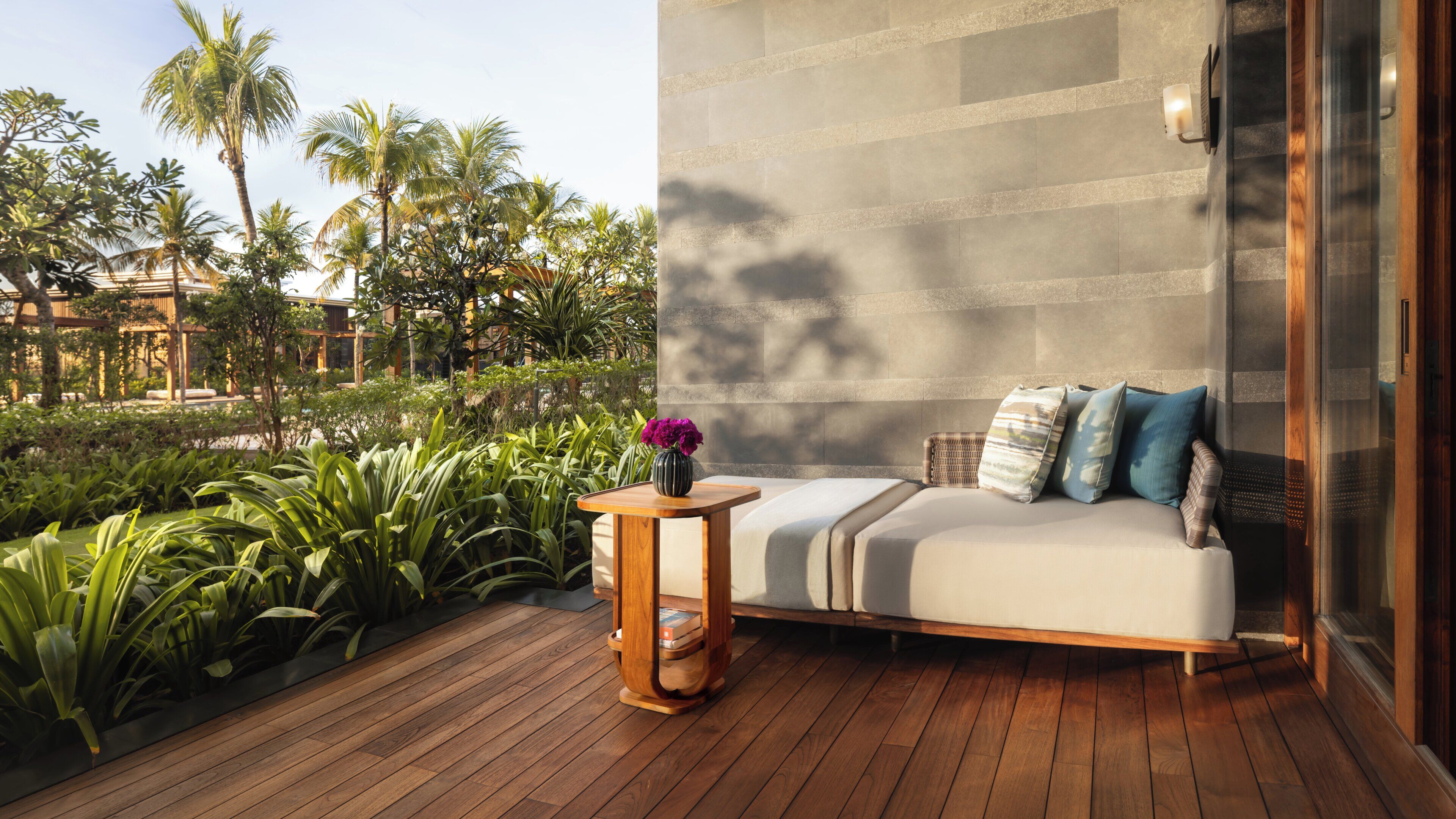 A modern outdoor seating area with a wooden deck, a cushioned sofa with pillows, and a vase of flowers.