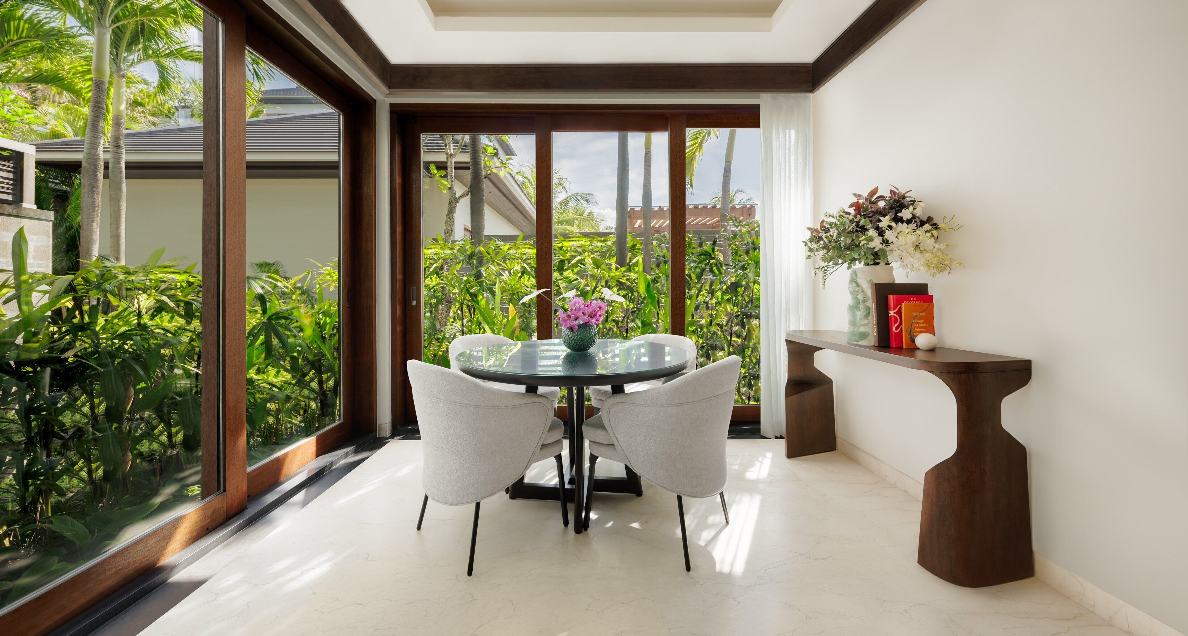 A dining area with a glass table and chairs, a vase with flowers, and a wooden sideboard.