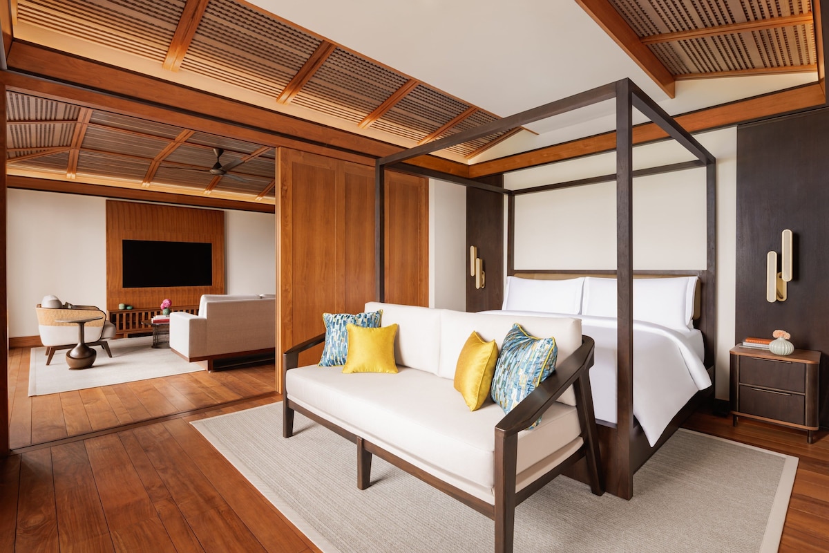 A spacious bedroom with a four-poster bed, a sofa, a TV mounted on the wall, and a wooden ceiling.