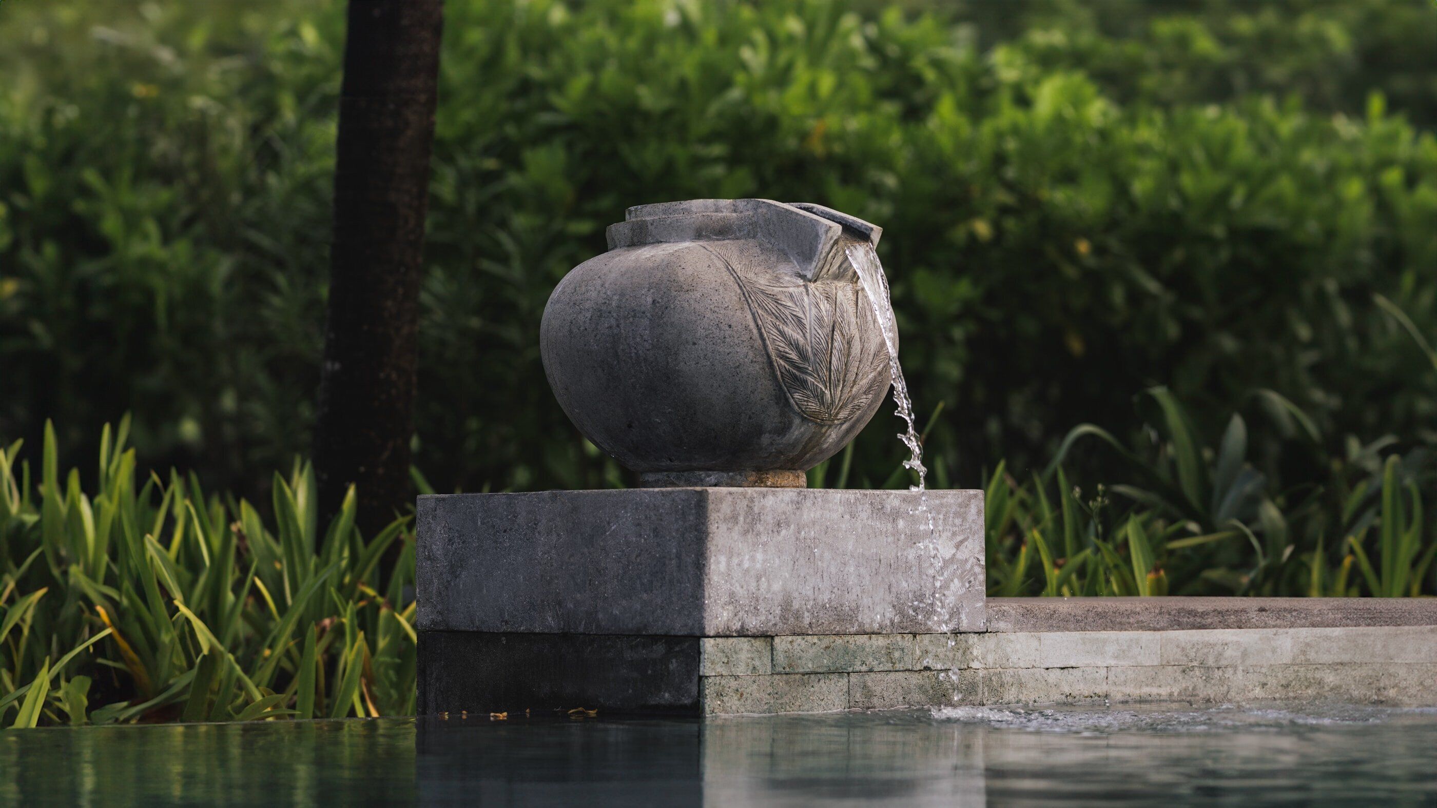 A stone water feature with a spout, set on a platform in a garden with lush greenery.