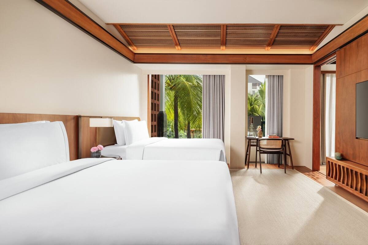 A hotel room with two beds, a TV, a small table with chairs, and a view of palm trees.