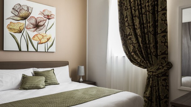 A hotel room with a bed, pillows, a lamp, a painting, and curtains. - Portside Lodge Building (Marsaskala)