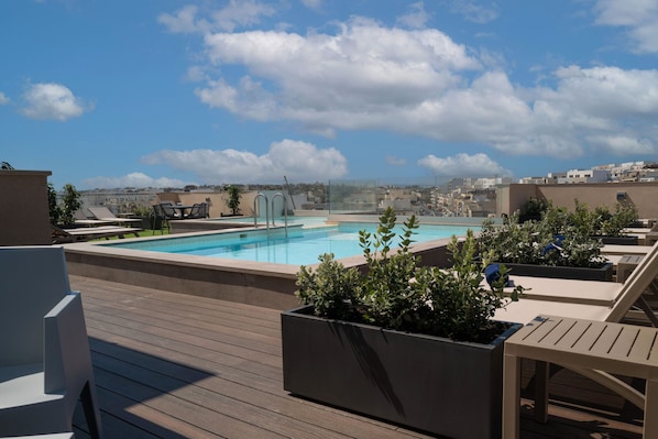 A rooftop pool with lounge chairs and planters, surrounded by buildings under a clear sky.