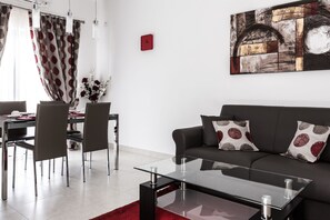 A modern living room with a glass coffee table, a dark sofa with patterned cushions, and a dining area with a table and chairs.