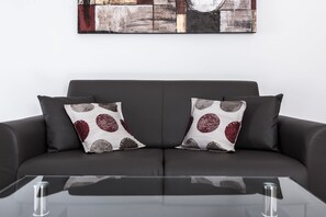 A modern living room with a dark grey sofa, two patterned throw pillows, and a clear glass coffee table. - Portside Lodge Building (Marsaskala)