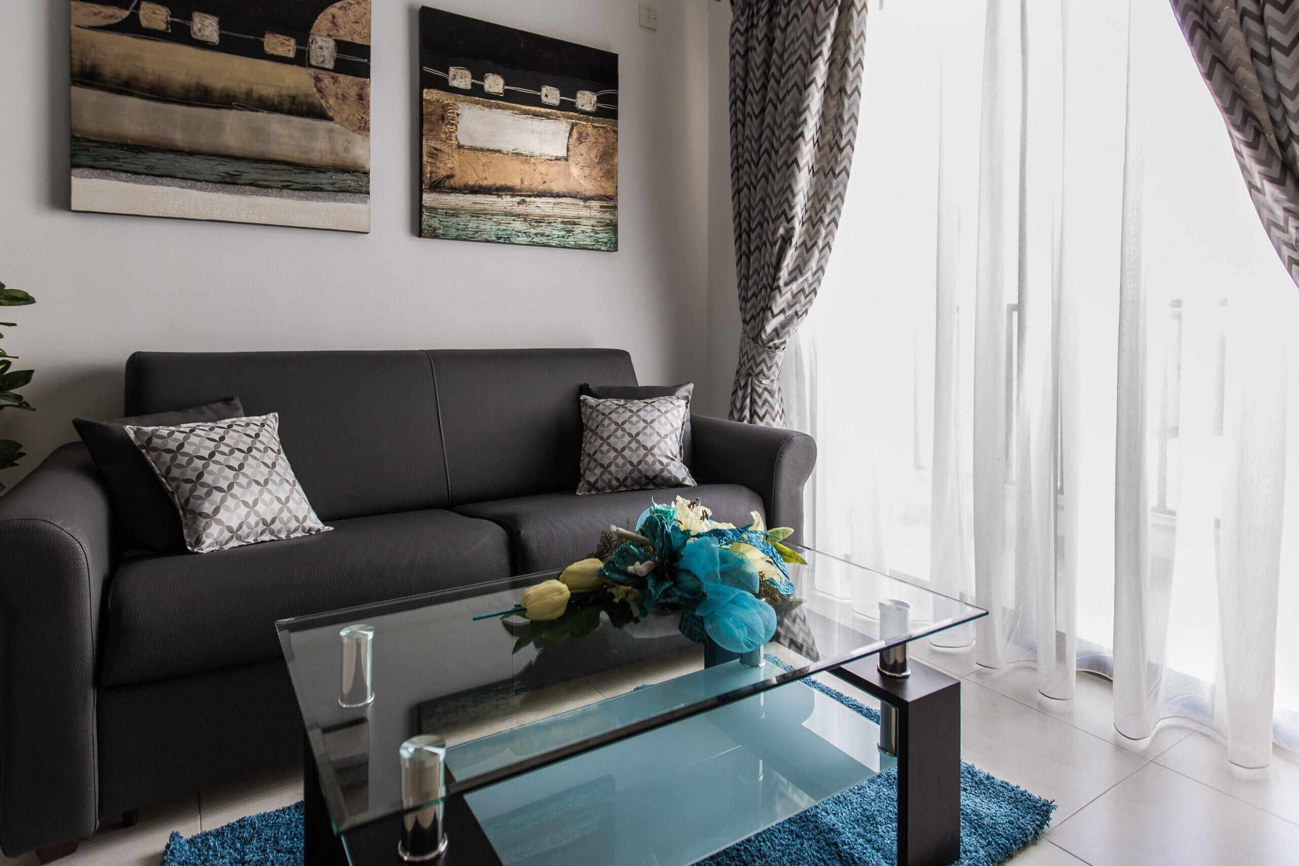 A modern living room with a dark sofa, patterned cushions, a glass coffee table with a floral arrangement, and abstract wall art.