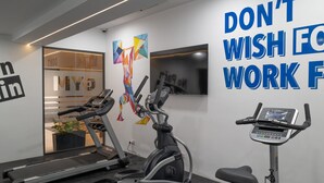 Ein Fitnessraum mit Laufbändern, Crosstrainern und einem Heimtrainer. An der Wand hängt ein Motivationsposter und im Hintergrund steht eine Pflanze.