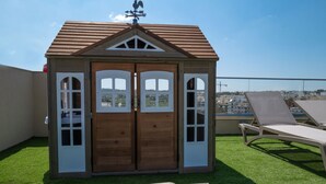 A wooden playhouse with a sloped roof and a weather vane on top, situated on a rooftop with artificial grass and a lounge chair. - Portside Lodge Building (Marsaskala)
