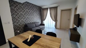 A modern living room with a grey sofa, a wooden dining table with a black placemat, and a TV mounted on the wall. - Portside Lodge Building (Marsaskala)
