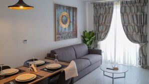 A modern living room with a dining area, a grey sofa, a coffee table, and a large window with curtains. - Portside Lodge Building (Marsaskala)
