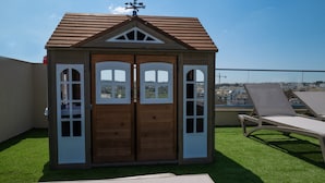 A wooden playhouse with a brown roof and white trim, featuring two doors and windows, situated on a rooftop with artificial grass and a view of the cityscape. - Portside Lodge Building (Marsaskala)