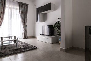 A modern living room with a flat-screen TV on a white cabinet, a glass coffee table, a grey rug, and a potted plant.