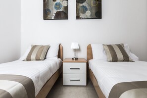 A hotel room with two single beds, a nightstand with a lamp, and a drawer unit.