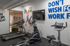 Ein Fitnessraum mit Laufbändern, Crosstrainern und einem Heimtrainer. An der Wand hängt ein Motivationsposter und im Hintergrund steht eine Pflanze.