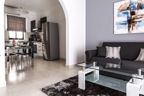 A modern living room with a glass coffee table, a black sofa with patterned cushions, and a contemporary kitchen in the background.