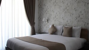 Cama bem arrumada com manta marrom, mesa de cabeceira com abajur e janela com cortinas.