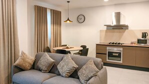 A modern living room with a grey sofa, a dining table, and a kitchen area with a stove and microwave. - Portside Lodge Building (Marsaskala)