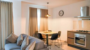 A modern living room with a grey sofa, a dining table with chairs, a kitchen area with a stove and oven, and a clock on the wall. - Portside Lodge Building (Marsaskala)