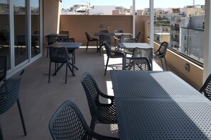 An outdoor patio with tables and chairs, large windows, and a view of the cityscape. - Portside Lodge Building (Marsaskala)