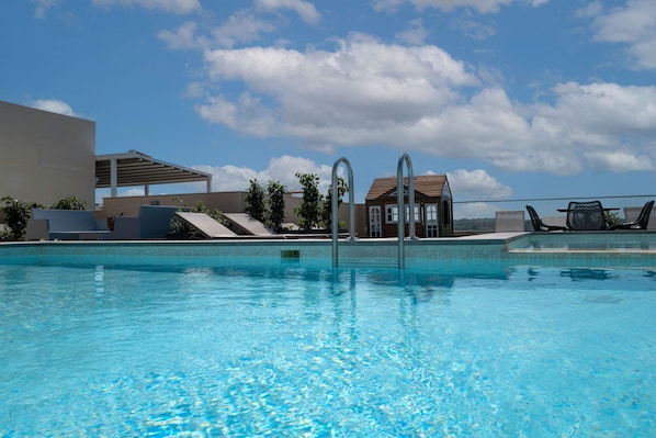 A swimming pool with clear water, lounge chairs, and a building with a tiled roof in the background.