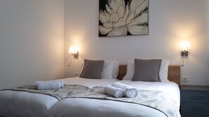 A neatly made bed with white linens and two rolled towels, a wall-mounted abstract painting, and bedside lamps on either side. - Portside Lodge Building (Marsaskala)