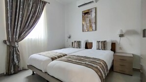 A double bed with a striped blanket, a bedside table with a lamp, and a painting on the wall. - Portside Lodge Building (Marsaskala)