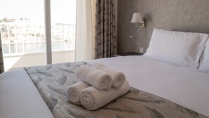 A hotel room with a bed, a view of buildings through a window, and a lamp on the wall. - Portside Lodge Building (Marsaskala)