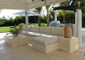 Relax in a shaded outdoor space with a sofa, ottoman, and woven baskets, near a grill and surrounded by lush greenery.