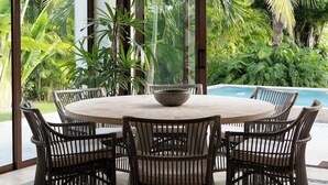 Een eethoek met een ronde tafel en rieten stoelen, een hanglamp en uitzicht op een zwembad en tropische planten.