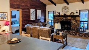 Un accogliente interno di una baita con camino in pietra, travi in legno a soffitto, zona pranzo e soggiorno con divano e mensola del camino.