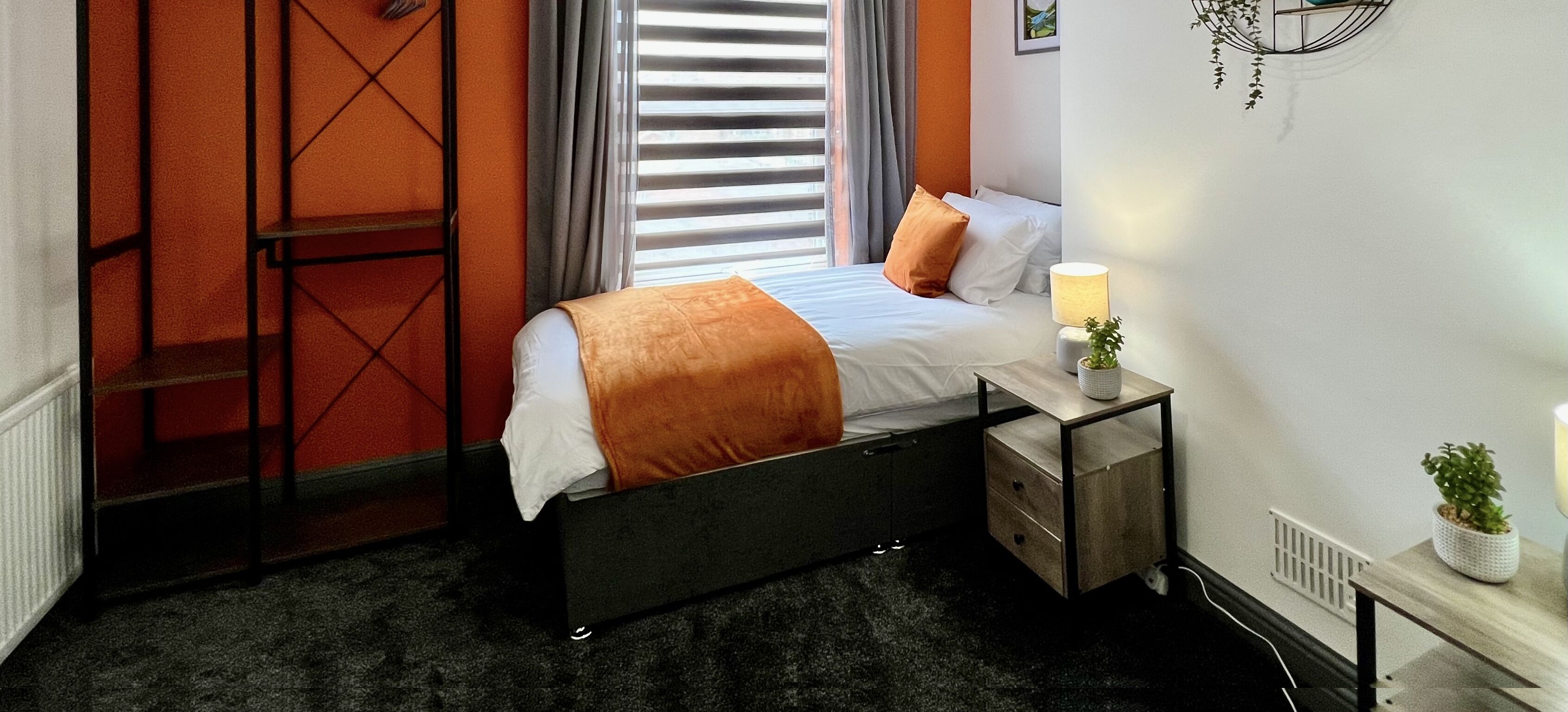 A modern hotel room with a bed, a bedside table with a lamp, a small plant, and a radiator.