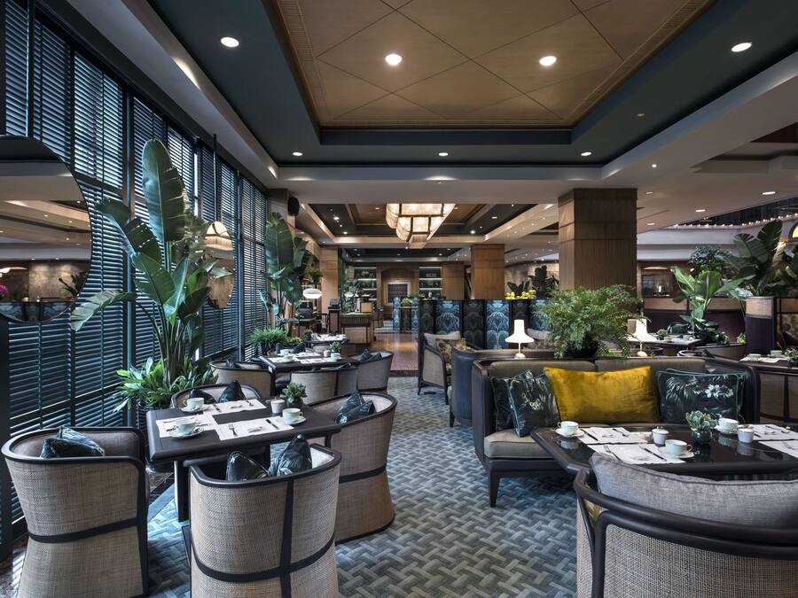 A spacious indoor area with multiple seating arrangements, including round tables and sofas, surrounded by potted plants and large windows with blinds.