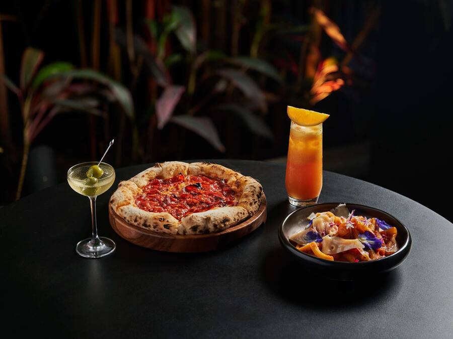 Une pizza sur une planche en bois, un verre à martini avec une tranche de citron et un saladier rempli de salade sur une table.