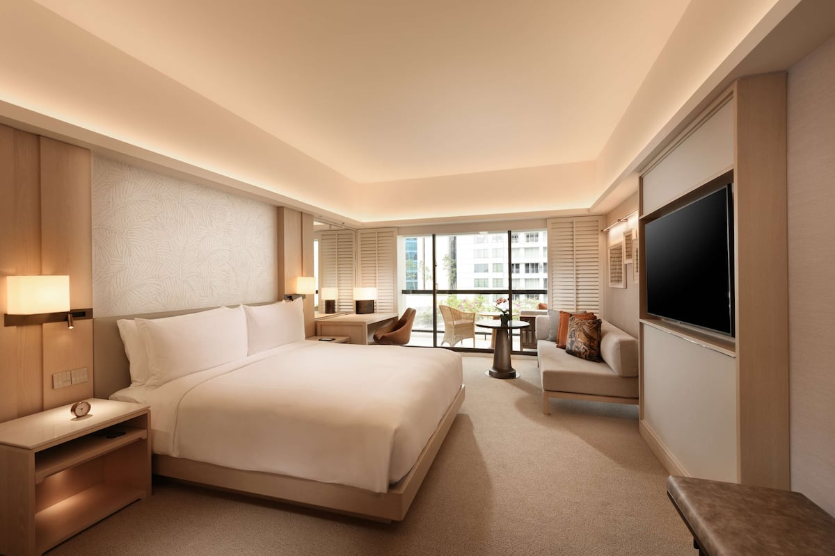 Een hotelkamer met een groot bed, nachtkastjes, een televisie, een zithoek met een tafel en stoelen, en een uitzicht op het exterieur door grote ramen.