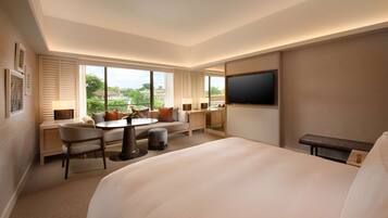 A hotel room with a large bed, a desk with a chair, a sofa, a TV, and large windows offering a view of the outdoors.