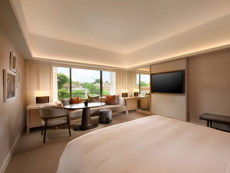 A hotel room with a large bed, a desk with a chair, a sofa, a TV, and large windows offering a view of the outdoors.