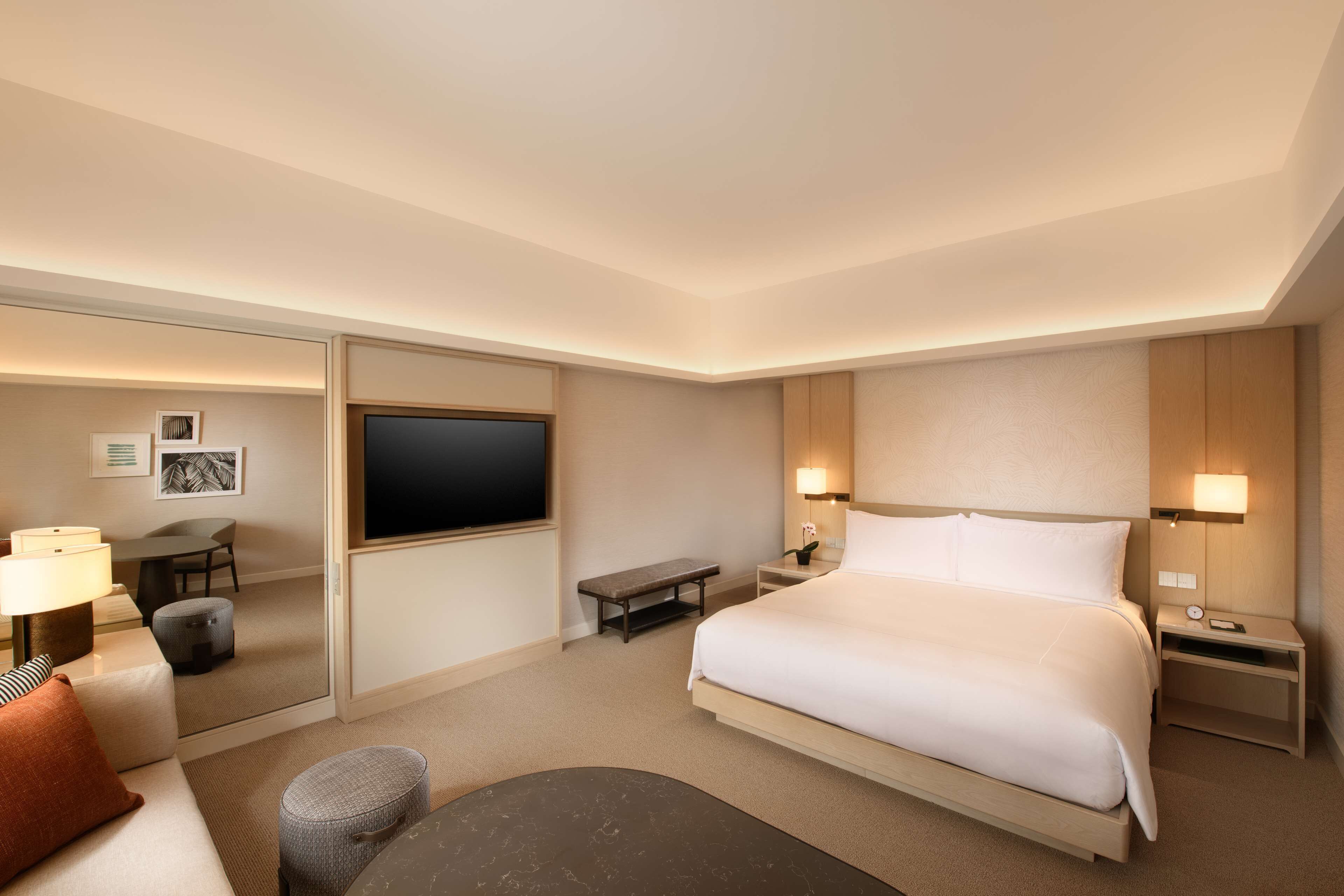 Habitación de hotel con una cama grande, mesitas de noche, un escritorio con una silla, un televisor y una zona de estar con un banco y un reposapiés.