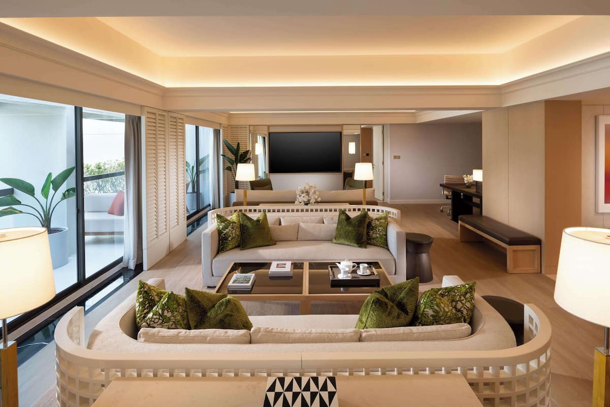 Een moderne woonkamer met een gebogen bank, een salontafel en een flatscreen televisie. Er zijn grote ramen met jaloezieën en potplanten.