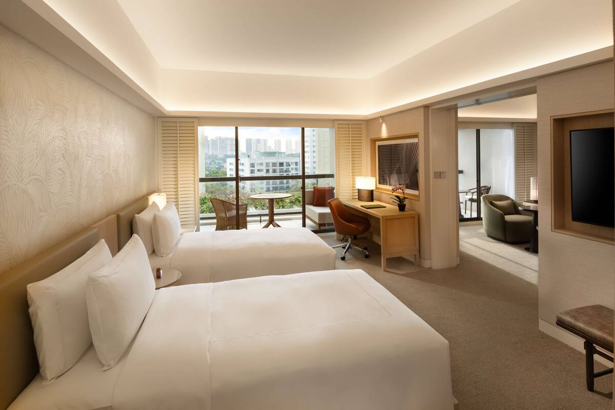 Een hotelkamer met twee bedden, een bureau, een stoel, een televisie en een balkon met uitzicht op de stad.