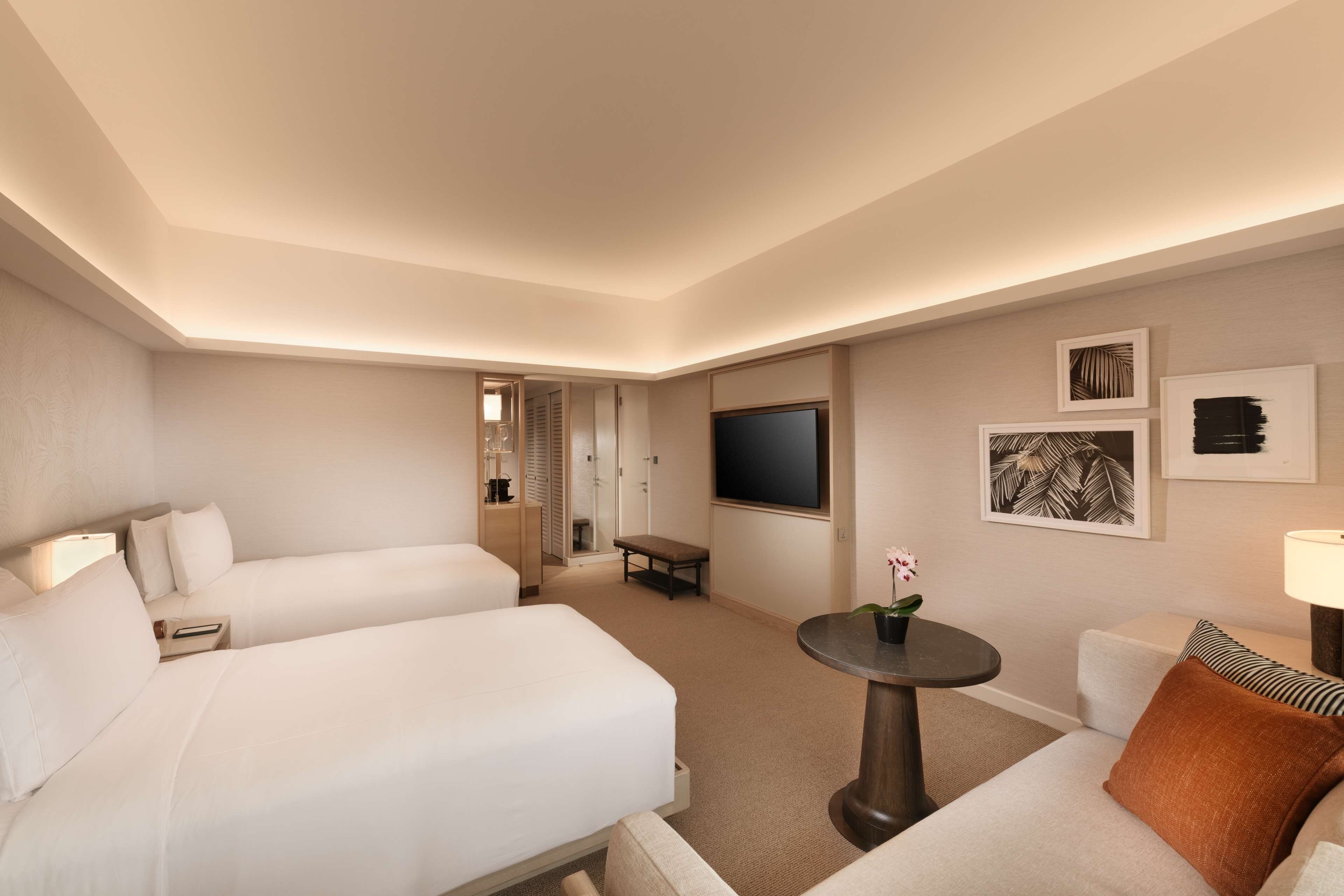 A hotel room with two beds, a TV, a small table with a flower, and a sofa with a pillow.