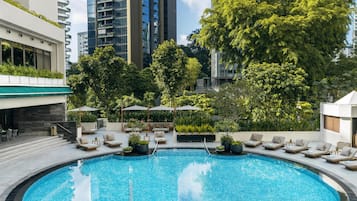 Piscina con sillas de descanso y sombrillas, rodeada de vegetación y edificios altos.