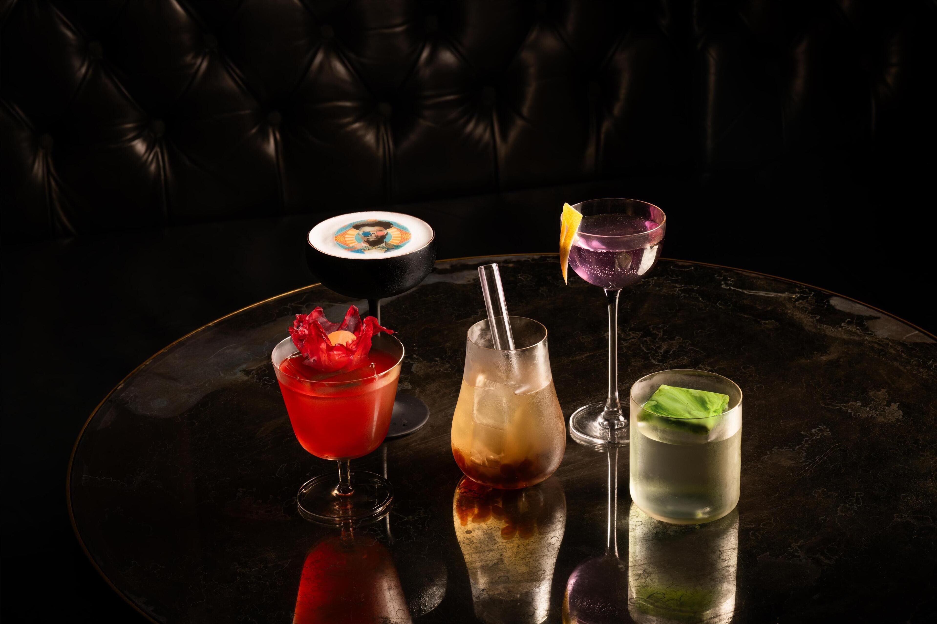 Una mesa redonda con cuatro bebidas distintas, un cuenco negro con diseño colorido y un sofá de cuero oscuro al fondo.